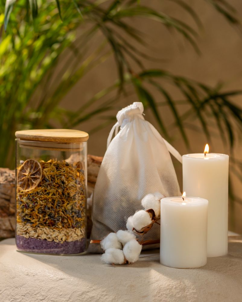 On a table, a jar of herbs, a candle, and cotton bag full of berbs are presented, symbolizing a peaceful environment for rejuvenating bath experiences.